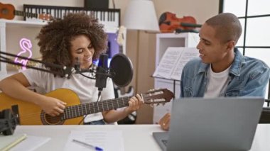 İki gülümseyen müzisyen rahat bir stüdyoda klasik bir gitarın klasik akortlarını tıngırdatıyor, keyifle şarkı söylüyor ve uyum sağlıyorlar, müzik mükemmelliği için bir dizüstü bilgisayar kullanıyorlar..