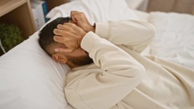 Süveter giymiş sakallı bir adam yatak odasında yorgun ve dinleniyor, sağlık, rahatlama ve yaşam tarzını tasvir ediyor.