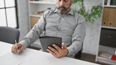 Tişört giymiş, gri saçlı, olgun sakallı bir adam modern ofis ortamında tabletine odaklanmış durumda..