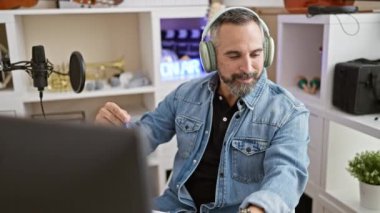 Ev stüdyosunda mikrofon ve kulaklık takan olgun sakallı bir adam..