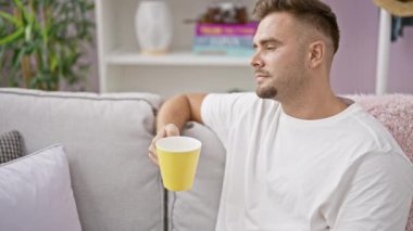 Rahat genç bir adam, elinde kupa tutarken rahat bir şekilde kanepede oturur, rahatlık ve sükunet hissi yayar..