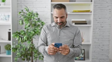 Gülümseyen, sakallı, gri saçlı, modern ofis ortamında akıllı telefon kullanan olgun bir adam..