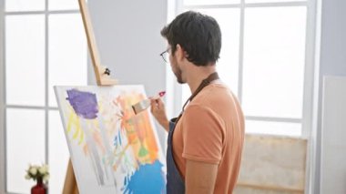 Parlak bir stüdyoda tuvale resim yapan sakallı bir adam. Gözlük ve önlük takıyor. Yaratıcılık ve odaklanma yayıyor..