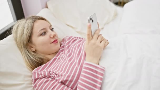若い金髪の女性は快適なストライプパジャマでスマートフォンを使用して笑顔で彼女の寝室でリラックス — ストック動画