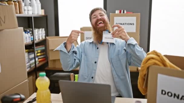 チャリティーセンターでボランティアをしている笑顔の赤毛の男は ノートパソコンで作業している間 彼の身分証明書を本気で指摘します — ストック動画