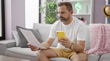 Orta yaşlı, gri saçlı, akıllı telefon kullanan ve modern oturma odasındaki kanepede kitap okuyan bir adam.