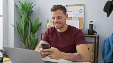 Modern ofis ortamında akıllı telefon kullanan sakallı yakışıklı genç adam rahat ve gülümsüyor..