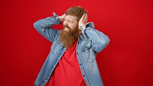 ストレスを受けた若い赤毛の男は 感情的な頭痛の問題を表現する赤い背景で孤立した騒々しい不安に耳を覆う疲れました — ストック動画