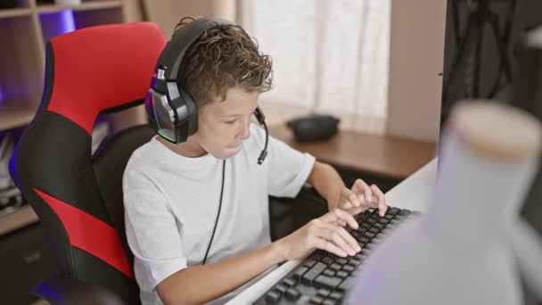 快適なブロンドの少年ストリーマーは 幸せにゲームをプレイし 将来的なゲームルームで自宅で自分のコンピュータ上でビデオゲームをストリーミングし 自信を持ってヘッドセットとゲームパッドを使用しています — ストック動画