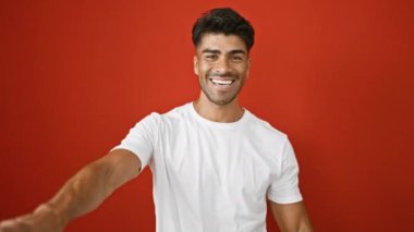 Beyaz gömlek giyen sakallı neşeli genç bir adam kırmızı duvar arkasında sanal bir sarılma hareketi yapıyor..