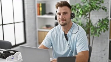 Kulaklık takan gülümseyen adam modern ofis içinde dizüstü bilgisayarla çalışıyor.