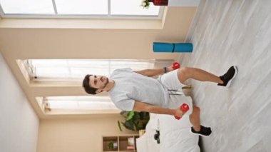 Parlak bir yatak odasında dambıllarla egzersiz yapan sağlıklı bir genç adam, ev içi çalışma rutinini gösteriyor..