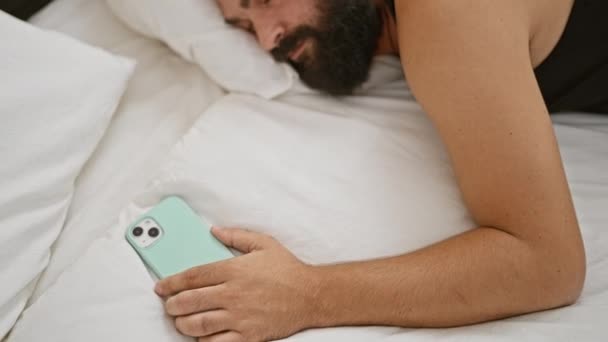 居心地の良い寝室のセットアップでスマートフォンで寝ているひげ付き男が現代的なライフスタイルのシーンをキャプチャします — ストック動画
