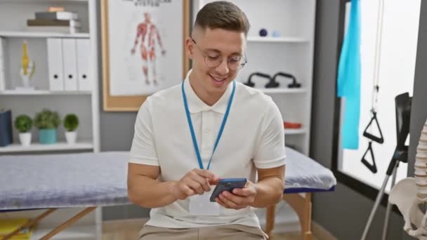 Χαμογελώντας Νεαρός Ισπανός Χρησιμοποιώντας Smartphone Στην Ιατρική Κλινική Απεικονίζει Επαγγελματία — Αρχείο Βίντεο
