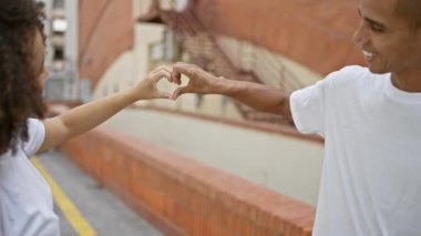 Güzel bir çift gülerek, güneşli bir kasaba caddesinde bir arada durarak, şık ve günlük yaşam tarzlarında mutluluk ve aşk yayarak kalp jesti oluşturuyorlar..