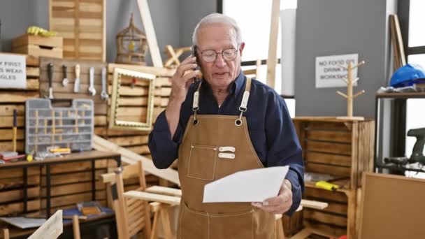 在他的木工车间里 一个成熟的男人弯着腰看文件 一边用智能手机聊天 一边在眼镜顶上下巴翘起 在他一生从事木工行业的中间交谈 — 图库视频影像