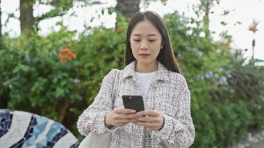 Çevresi yeşilliklerle çevrili, akıllı telefon kullanan Asyalı genç bir kadın.