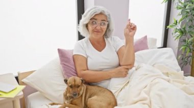 Gri saçlı köpeği olan neşeli orta yaşlı kadın, yan tarafa bakıyor ve gülümsüyor, rahat bir odada pijamada mutlu bir şekilde oturuyor.