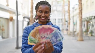Güney Afrika rand 'ını elinde tutan gülümseyen Afrikalı kadın güneşli bir şehir sokağında notlar tutuyor.