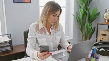 Profesyonel orta yaşlı bir kadın dizüstü bilgisayarda çalışır ve modern ofis ortamında akıllı telefonunu kontrol eder..
