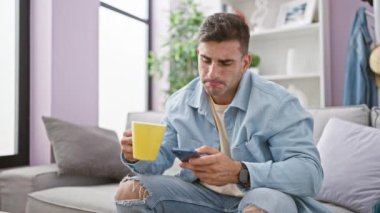 Zeki telefon kullanmaya dalmış, oturma odasındaki kanepede dinlenirken rahat evinde espresso içen çekici, genç bir İspanyol.