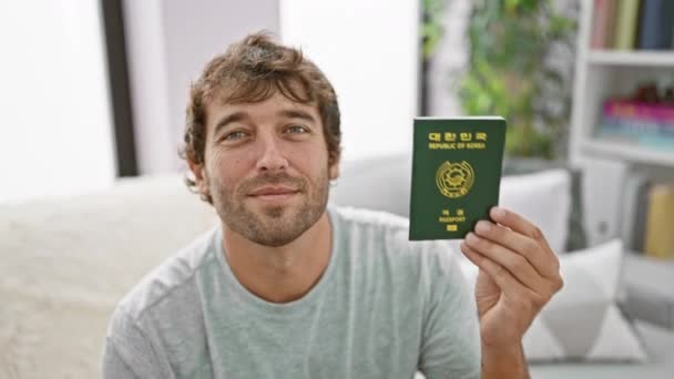 自宅で笑顔で韓国のパスポートを持っているソファーに座っている若い男 — ストック動画