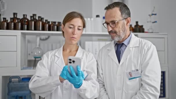 2人の献身的な科学者 男性と女性は 深刻な科学研究室内のスマートフォンで発見を話し 共有し エキサイティングな医学研究に密接に取り組んでいます — ストック動画