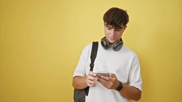 クールな若いヒスパニック系男性 ティーンエイジャー学生のロキン黄色いバックパック スマートフォンで音楽に詰め込み ヘッドフォンを着用し 大きな時間を祝い 黄色い壁に隔離された完全に静かな — ストック動画