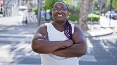 Afro-Amerikan kadın, spor örgüleri ve gözlükleriyle, geniş bir alanda gülümsüyor ve şehir caddesinde çapraz kollarla duruyor, dışarıda neşe ve ifade yayıyor..