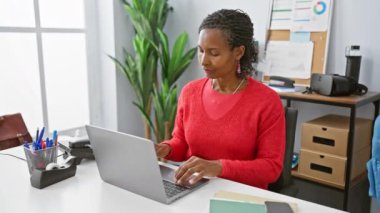 Kırmızı kazaklı olgun bir Afrikalı kadın ofis masasında dizüstü bilgisayar ve kırtasiye malzemesi ile meditasyon yapıyor..