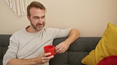 Kırmızı kupa tutan gülümseyen sakallı adam, rahat bir oturma odasındaki gri koltukta oturuyor.