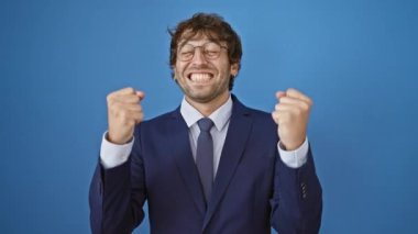 İş elbisesi içindeki neşeli genç adam, İspanyol ve sarışın, zafer kollarını kaldırışını kutluyor. Destansı galibiyet, başarıyla ışıldıyor! Mavi bir arkaplanda izole edilmiş.