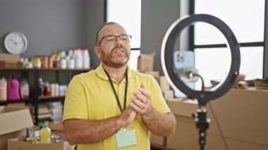 Çekici, orta yaşlı, sakallı beyaz bir adam, kendini işine adamış bir gönüllü olarak, bağışların ve kutuların arasında, hayır merkezi video görüşmeleri sırasında olumlu jest yapıyor.