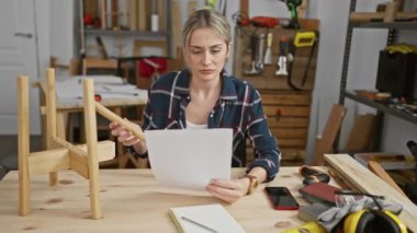 Aletler ve ahşap malzemelerle çevrili bir marangoz atölyesinde planlar okuyan odaklanmış bir kadın..