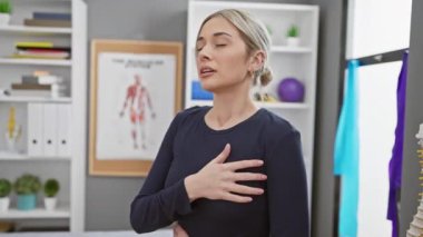 Klinikteki sarışın kadın göğsüne dokunuyor fizyoterapi ekipmanları ve anatomi posterleriyle..