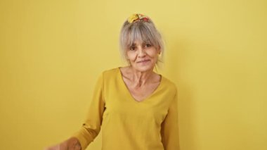 Midilli kuyruklu, beyaz saçlı neşeli orta yaşlı kadın mutlu bir gülümsemeyle parmaklarının arasından röntgencilik yapıyor. Parlak sarı bir arkaplanda izole edilmiş..