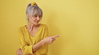 Kendine güvenen orta yaşlı, gri at kuyruklu bir kadın, izole edilmiş sarı bir arka planda ciddi bir yüzü olan yan reklamı işaret ediyor, güvenilir bir figür yolu gösteriyor.