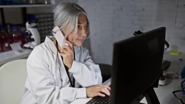 在下班后的实验室里 一位漂亮的 灰白头发的科学家在不停地工作 做着多项任务 在使用电脑时 她一边用智能手机一边说话 一边洋溢着中年人的钢铁般的决心 — 图库视频影像