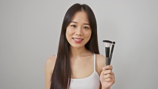喜びの若い中国の女性は自信を持ってメイクブラシを展示し 白い背景に隔離された静かな笑顔を輝かせます — ストック動画