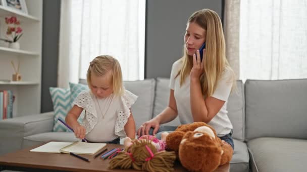 リラックスした注意深い母と小さな娘が自宅に座り ノートブックに絵を描いていました リビングルームテーブルのおもちゃ 人形の中でスマートフォンで笑顔の家族のチャット マルチタスク — ストック動画