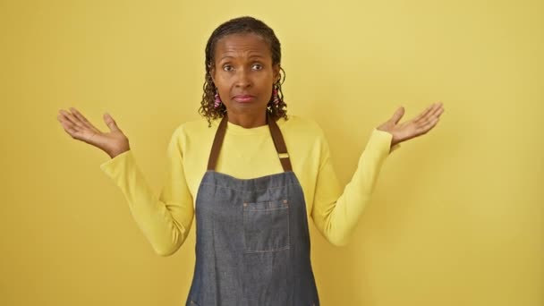 無力で混乱した美しい中年のアフリカ系アメリカ人女性エプロンクラッドは 孤立した黄色の背景に対して手を挙げ 疑いの完璧な画像をキャプチャします — ストック動画