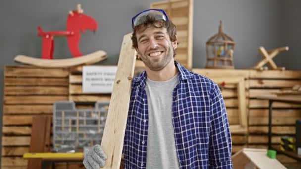 英俊的年轻人留着胡子 熟练的木匠 在他的行业里是个行家 在眼镜后面笑着 把木板放在木工车间里 准备好征服木材业 — 图库视频影像