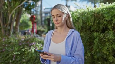 Çiçekli bir bahçede akıllı telefon kullanan gülümseyen sarışın bir kadın.