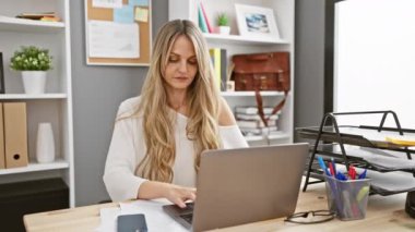 Modern ofis ortamında akıllı telefon ve dizüstü bilgisayarla çalışan odaklanmış sarışın bir kadın.