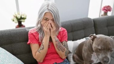 Duygusal tablo, gri saçlı orta yaşlı bir kadın, stresli ve endişeli, kanepeye oturmuş, gözyaşlarına boğulmuş bir köpekle, olgun yüzü endişe içinde, oturma odasında kendini iyi hissetmediğini ifade ediyor..