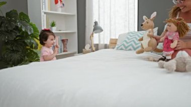 Kendine güvenen anne ve neşeli kız oyuncaklarla oyun oynuyorlar, güneşli bir yatak odasında rahat bir yatakta oturuyorlar, sıcak bir gülümsemeyle neşe ve sevgiyi ifade ediyorlar.