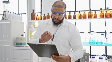 Laboratuvar önlüğü ve güvenlik gözlüğü takmış sakallı bir Afrikalı Amerikalı, şişelerle dolu bir laboratuvar ortamında notları inceliyor..