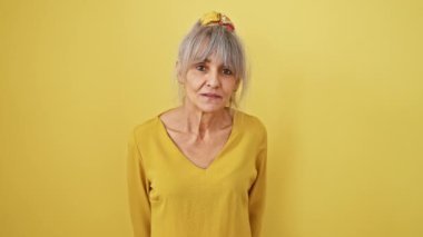 Beyaz saçlı ve at kuyruklu neşeli orta yaşlı kadın kameraya öpücük veriyor, özgüven ve seksilik fışkırıyor. Havada aşkı ifade etmek eğlenceli sarı bir arka planda.