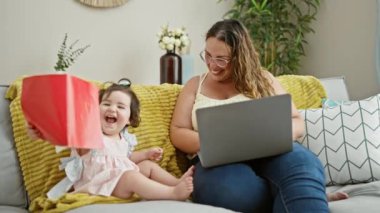 Gülümseyen anne ve kız rahat bir anın tadını çıkarıyor, oturma odasındaki koltuklarında oturuyor, dizüstü bilgisayarlarında interneti keşfediyor, iş ve aşkı dengeliyor, evde en sevdikleri kitabı tutarken