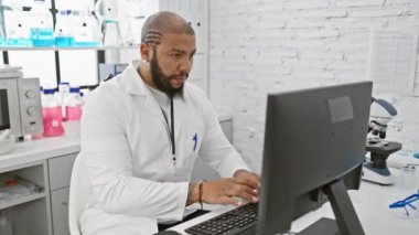 Focused black man in lab coat working on computer in modern laboratory setting